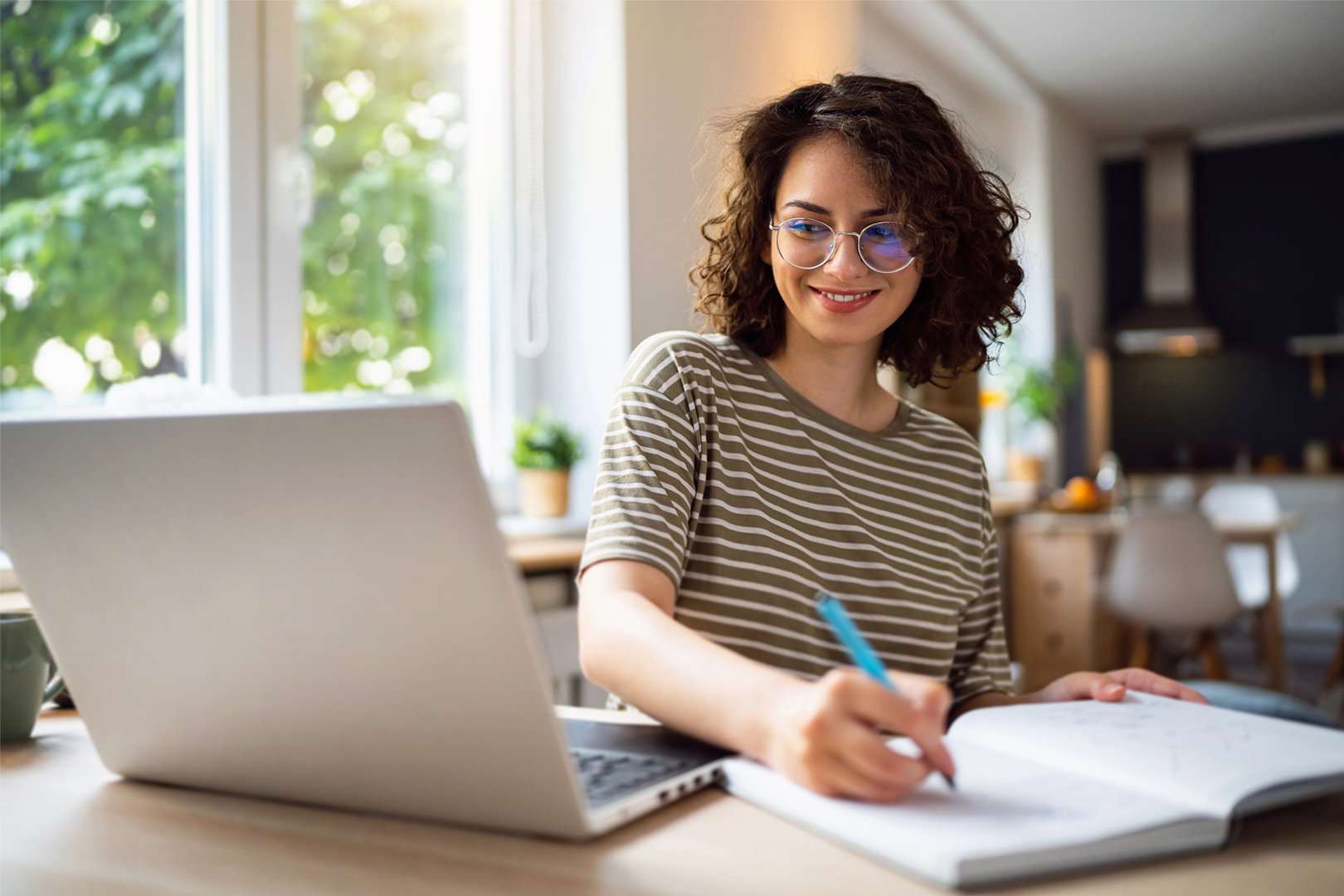 A woman seated at a desk looking at a computer while writing in a notebook