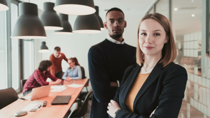 Confident young professionals standing in a modern office with coworkers collaborating in the background.